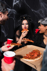 pleased asian woman in vampire halloween costume eating pizza near multiethnic friends on black