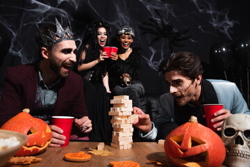 man in halloween makeup playing wood blocks game while blurred interracial women toasting with...