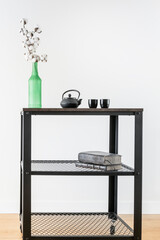 Industrial style trolley with green bottle with decorative cotton branch and black tea set with old books and white wall in the background