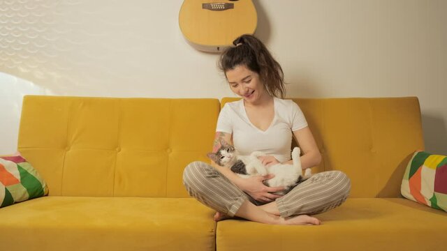 Strong and independent child free female spend her free time with pets. Young pretty woman sitting on coach at home with her cats. Girl playing and hugging adorable kitty. Animal lover concept.