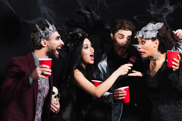 cheerful multiethnic friends with plastic cups singing karaoke during halloween party on black