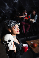 smiling african american woman in werewolf halloween mask showing spooky skull on black