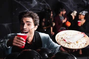 happy man in halloween makeup drinking beer near balloons and blurred friends on black background