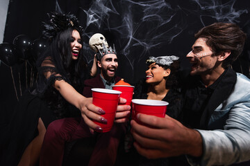 happy multiethnic friends in halloween costumes toasting with plastic cups on black