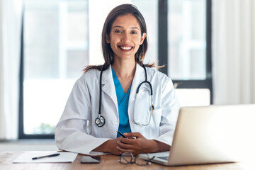 Attractive female doctor smiling looking at camera while working with laptop in the consultation.