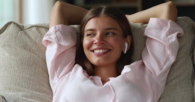 Tranquil gen z female teenager wear modern wireless headphones rest on soft cozy sofa engaged in easy learning english language audio course. Happy young lady take break relax lying hands behind head