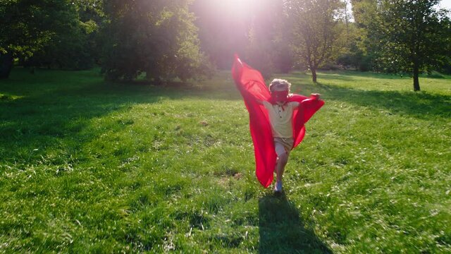 In a large green park running through the grass a cute small boy in a superhero suit he are very excited and happy. 4k