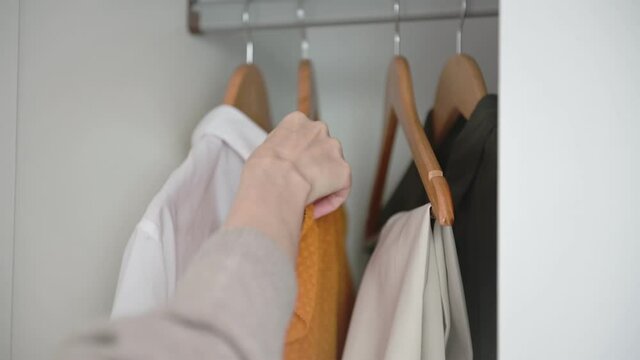 Woman hand opens white wardrobe with colorful clothes chooses and takes short-sleeved orange dress and closes closet closeup