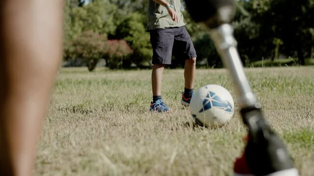 Close-up of mans artificial leg kicking ball around. Disabled father training, playing football with son on lawn in summer park. Disability, sport, outdoor activity concept