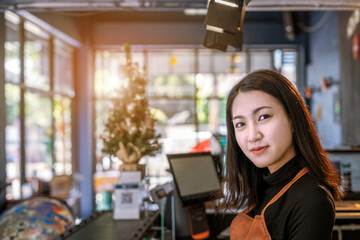 Portrait of confident Asian young adult owner gen z young woman at a counter bar in coffee shop cafe. She is wearing an apron looking at the camera and smiling. Hospitality and welcome concept