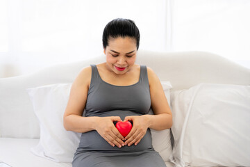 Pregnant woman holding red heart touching her belly. Loving mom waiting of a baby. Maternity, parenting, prepare and expect concept. Focus red heart