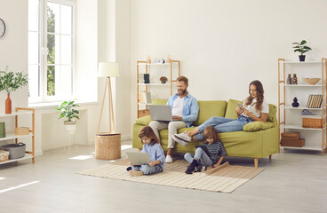 Family sitting in cozy spacious Scandinavian livingroom with wireless Internet connection at home, kids watching video on laptop and tablet, parents using their own devices. People, technology concept