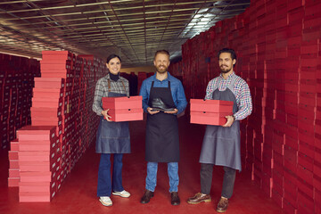 Team of happy proud smiling factory workers with red cardboard boxes showing brand new leather boot...