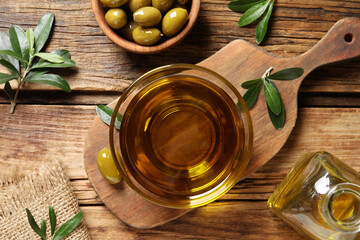 Glass bowl of fresh oil, ripe olives and green leaves on wooden table, flat lay
