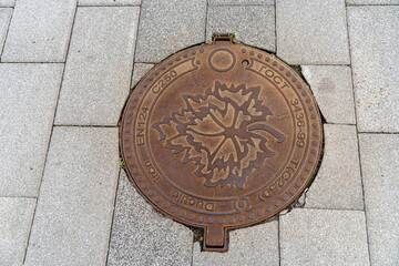 A cast iron manhole cover with embossed maple leaf is positioned on the gray concrete slab sidewalk.