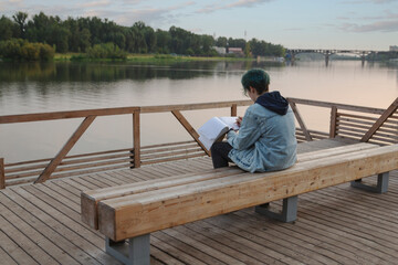 Girl draws in a sketchbook while sitting on the embankment in summer