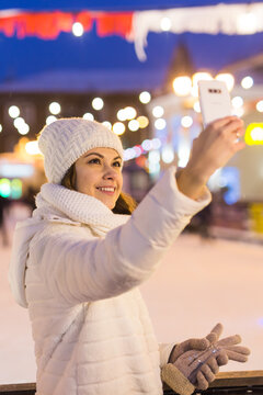 Christmas, winter, technology and leisure concept - happy young woman taking picture with smartphone on ice skating rink outdoors.