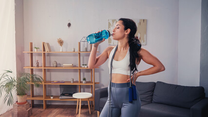 Side view of sportswoman with jump rope drinking water at home - obrazy, fototapety, plakaty