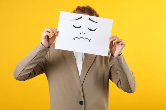 Woman hiding behind sheet of paper with sad face on yellow background