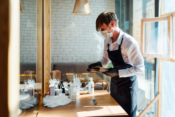 A joyful waiter in a mask stands near the table with personal hygiene products and disposable utensils. Precautions during quarantine in a cafe.