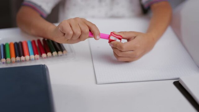 Close up of kid sitting at desk and using sharpener for sharpening colorful pencils. Little child preparing tools for drawing. Creative time spending at home.