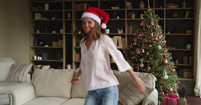 It is Christmas. Active gen z female dance by music celebrate winter holidays in cozy modern living room with decorated fir tree. Energetic young woman having fun enjoy New Year party for one at home