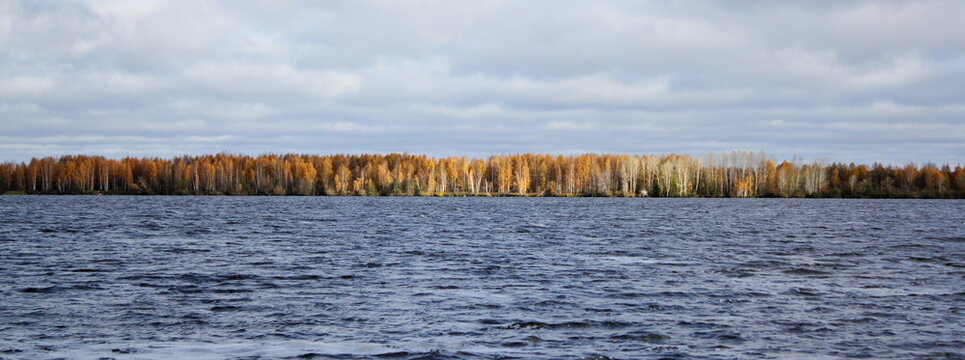 Photo of the Russian landscape. It depicts a large river and a bank with a green forest. Time of day is morning, the rays of the sun illuminate the forest on the other side of the river.