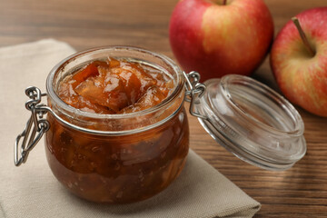 Tasty apple jam in glass jar and fresh fruits on wooden table, closeup