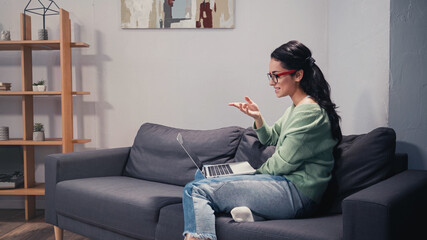 Obraz premium Side view of smiling freelancer having video call on laptop on couch