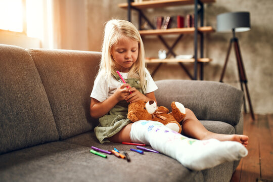 Little girl with a broken leg on the couch. The child draws with felt-tip pens on a plaster bandage.