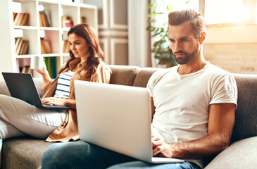 Young couple in love are sitting on the couch with laptops in the living room at home. Online shopping, work from home, freelance.