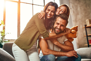 Happy dad and mom with their cute daughter and teddy bear hug and have fun sitting on the sofa in...