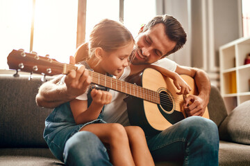 Happy dad teaches his cute daughter to play the guitar while sitting on the sofa in the living room...