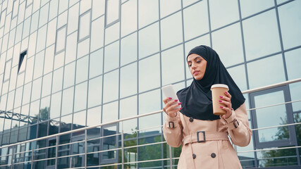 young muslim woman in hijab using smartphone and holding coffee to go near building