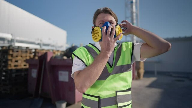 Portrait of brunette Caucasian adult man putting on respirator mask in slow motion looking around standing at sunny warehouse storage outdoors. Confident expert at workplace. Industry and protection