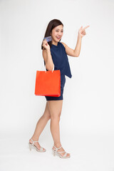 Portrait of an excited beautiful woman holding red shopping bags and credit card and pointing finger to empty space isolated over white background, Asian model and full body composition