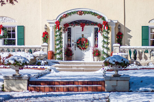 Upscale Landscaped  Stucco House With Elegant Entryway With Columns Decorated For Christmas With Big Wreath And Garland On Bright Snowing December Day