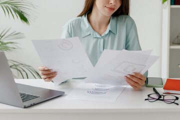 Cropped young successful employee business woman 20s in blue shirt hold paper account documents sit...