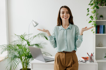 Young fun smiling successful employee business woman in casual blue shirt work look camera stand spread hands near workplace desk with laptop pc computer at office indoors. Achievement career concept.