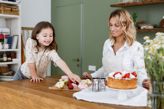 Happy chef cook baker mom woman in white shirt work with child baby girl helper take food from kitchen table at home. Cooking food process concept Mommy little kid daughter prepare fruit sweet cake