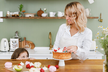 Fun chef cook baker mom woman in white shirt work with baby girl helper hiding behind table at kitchen table home. Cooking food process concept Mommy little kid daughter prepare fruit sweet pie cake.