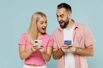 Young surprised happy couple two friends family man woman in casual clothes holding use mobile cell phone look to each other together isolated on pastel plain light blue background studio portrait