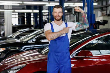 Strong young technician car mechanic man in blue overalls white t-shirt show point on biceps...
