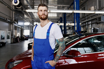 Strong pensive young male professional technician car mechanic repairman man 20s in denim blue overalls white t-shirt look aside work in modern vehicle repair shop workshop inside Tattoo translate fun