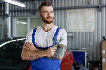 Minded young technician car mechanic repairman man in denim blue overalls white t-shirt hold wrench...