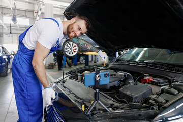 Young technician car mechanic man in blue overalls white t-shirt talk mobile cell on phone stand show troubleshooting process fix problem with raised hood work in vehicle repair shop workshop indoors - obrazy, fototapety, plakaty