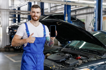 Young technician car mechanic man in blue overalls t-shirt use hold mobile cell phone blank screen...