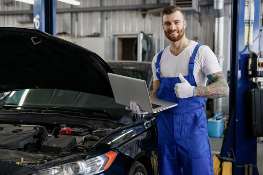 Young technician car mechanic man in blue overalls white t-shirt use hold laptop pc computer make diagnostics show thumb up fix problem with raised hood work in vehicle repair shop workshop indoor.