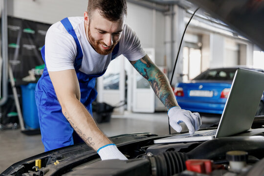 Young fun happy professional technician car mechanic man in blue overalls t-shirt use laptop pc computer make diagnostics check fix problem with raised hood work in vehicle repair shop workshop inside