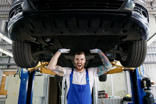 Strong excited young male professional technician mechanic man wears denim blue overalls white t-shirt stand near car lift posing like carry automobile work in vehicle repair shop workshop indoors